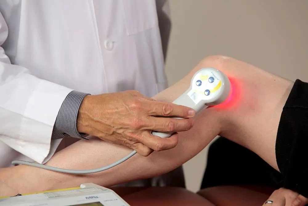 A healthcare professional uses a medical device emitting red light on a patient's knee.