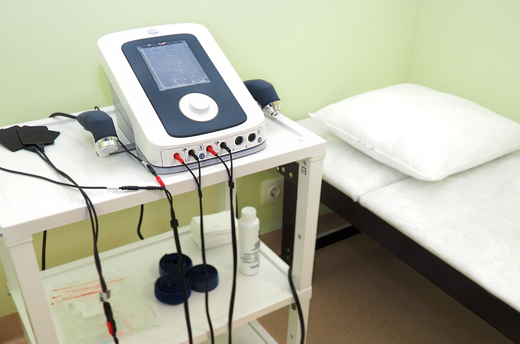 A medical device on a cart with various cables and accessories next to a cushioned examination bed.